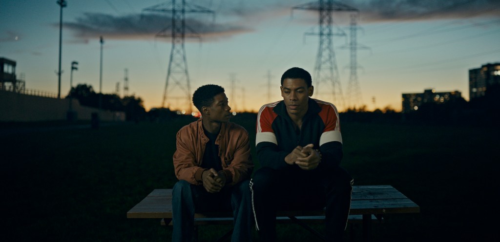 Two young Black men sitting on a wooden bench in an outdoor park at dusk, with power lines in the background and urban buildings faintly visible in the distance.