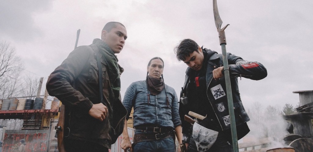 Three young Indigenous men stand together, dressed in rugged clothing, in a gritty outdoor setting. One holds a spear while another appears to be adjusting his gear. The scene suggests tension or conflict, reflecting themes of resilience and identity. Film is Jeff Barnaby's Blood Quantum