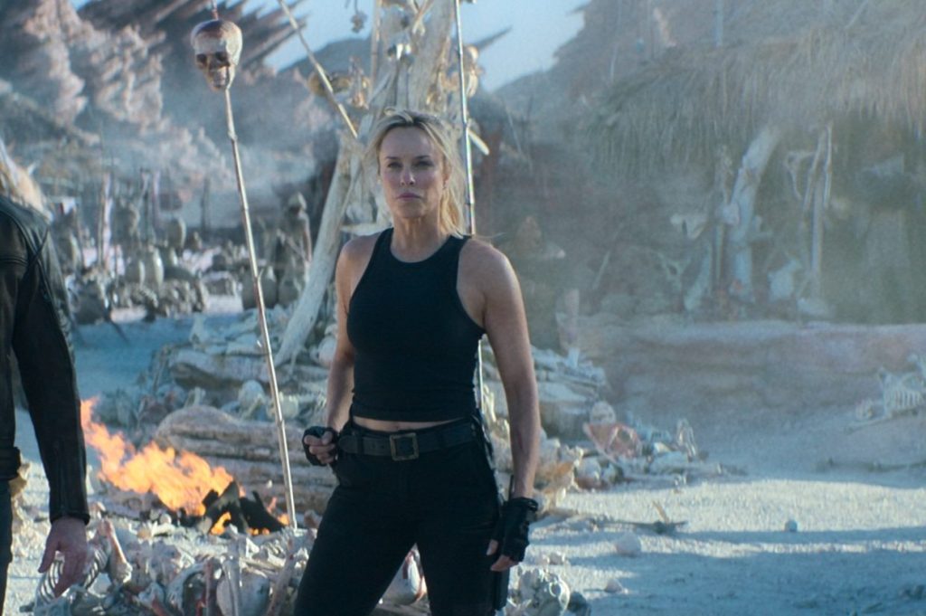 A woman in a black tank top and tactical pants stands confidently in a desolate landscape filled with skeletal remains and scattered debris, with a fire burning nearby.