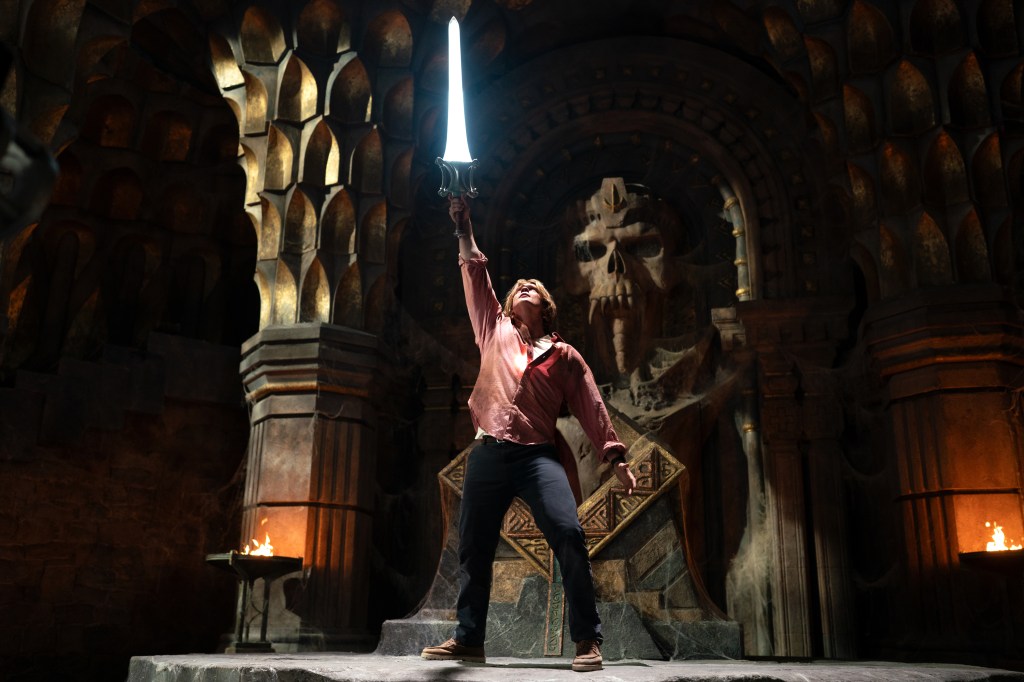 A character triumphantly raises a glowing sword in an ancient, dimly lit chamber featuring stone sculptures and flickering flames.