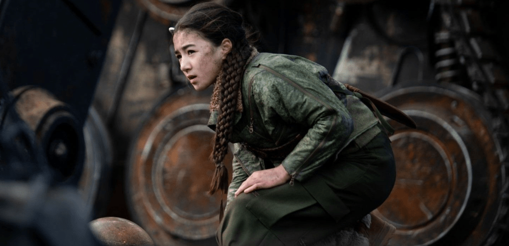 Young girl in green military-style clothing, crouched down with a serious expression, against a backdrop of industrial machinery.