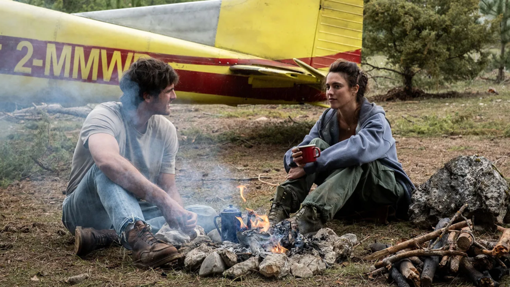 A man and a woman sit by a campfire in a forested area, with a small airplane in the background. The man is wearing a light shirt and jeans, while the woman is dressed in a sweater and cargo pants, holding a red cup.