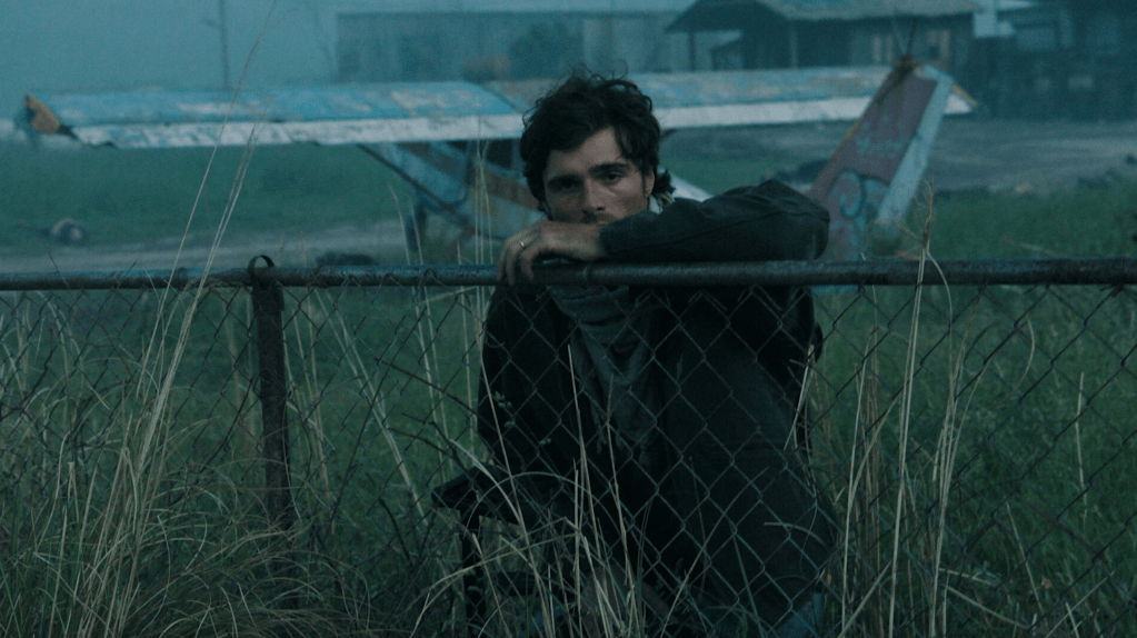 A young man leans against a chain-link fence in a foggy setting, with an abandoned airplane in the background and tall grass surrounding him.